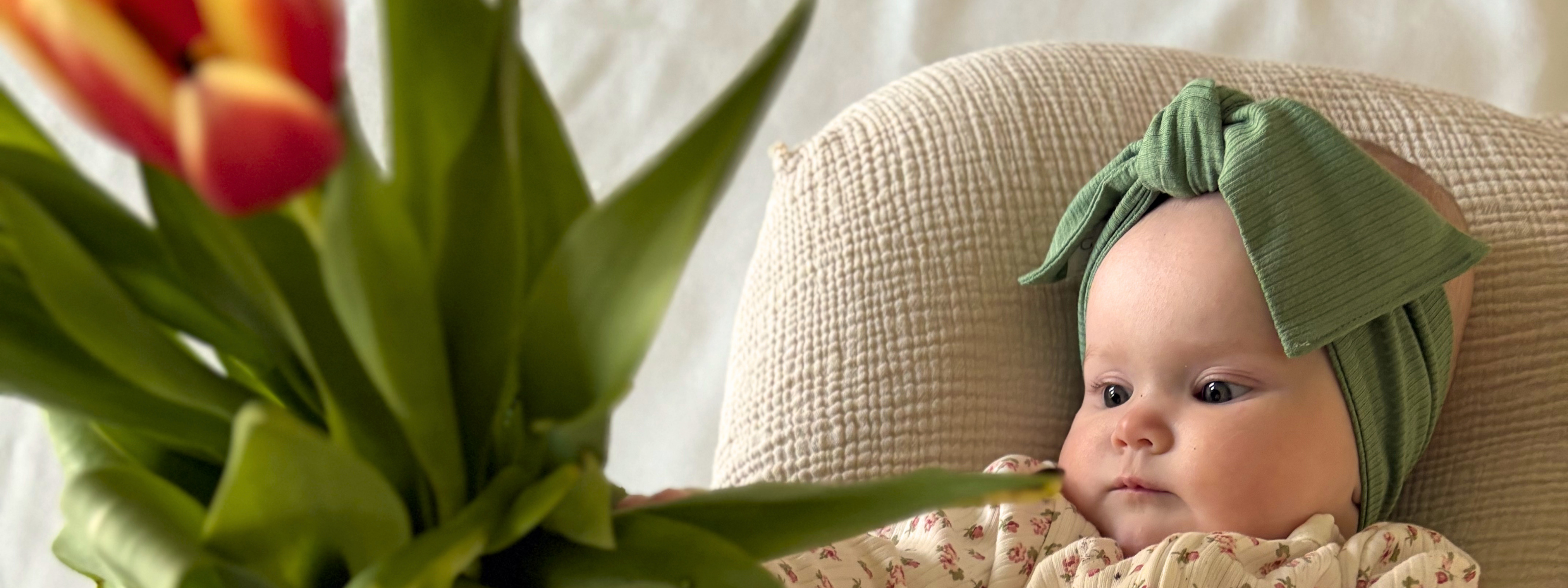 Baby wearing a green headband sitting on a cushion next to a vase of tulips.